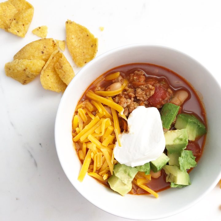 a white bowl filled with chili topped with cheddar cheese, sour cream, and avocado with corn chips on the side.
